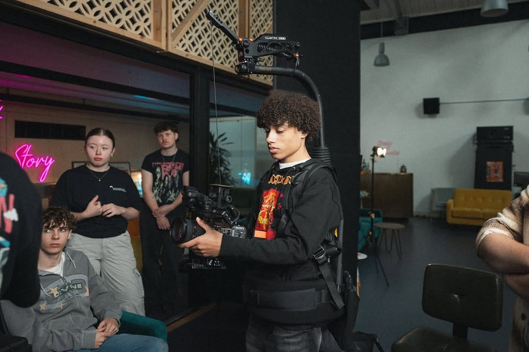 A filmmaker training on a steadycam in a large room with other trainees.
