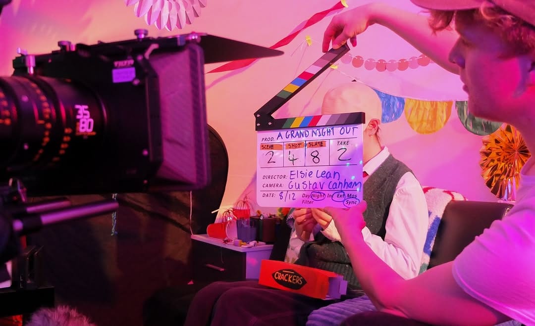 A filmmaker holds a clapper board in front of a camera in a pink room with decorations.